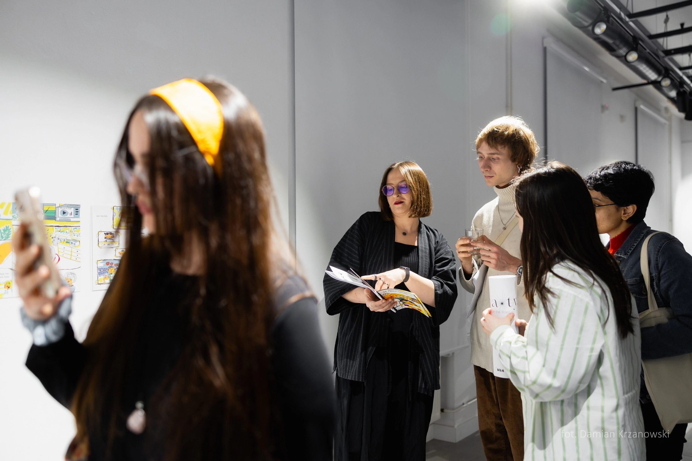 Grupa kilku osób stoi w przestrzeni galerii sztuki podczas wernisażu wystawy. W centrum kobieta - Agata Sztorc-Gromaszek trzyma publikację towarzyszącą wystawie i rozmawia z kilkoma osobami, pokazując coś w tekście broszury. Jedna osoba trzyma szklankę napoju. Na pierwszym planie, lekko rozmyta, znajduje się kobieta z długimi włosami i żółtą opaską, robiąca zdjęcie telefonem. Oświetlenie jest jasne, punktowe. 