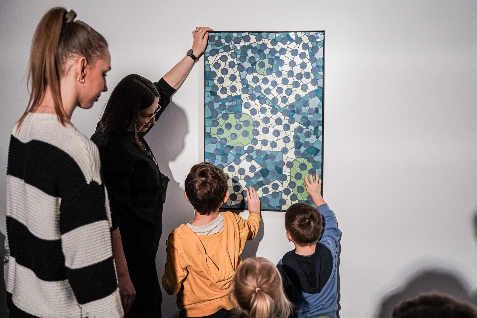 Grupa osób w galerii sztuki ogląda obraz zawieszony na ścianie. Dwie kobiety towarzyszą trojgu dzieciom, które stoją blisko pracy i dotykają jej powierzchni. Jedna z kobiet podtrzymuje ramę obrazu, a druga obserwuje z boku. Dzieło ma abstrakcyjny, geometryczny wzór złożony z połączonych kształtów w odcieniach niebieskiego, zieleni i bieli. Fotografię wykonano podczas warsztatów lub zajęcia edukacyjnych.